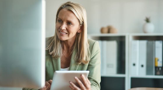 Eine Frau blickt in ihrem Büro auf den Desktop ihres Computers sowie auf das Display ihres Tablets.