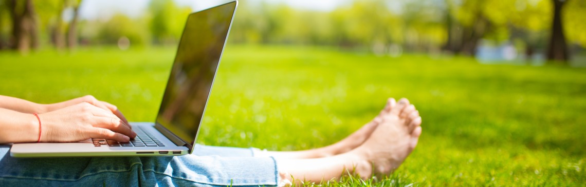 Eine Frau sitzt barfüßig und mit einem Laptop auf ihrem Schoß in einer grünen Wiese.