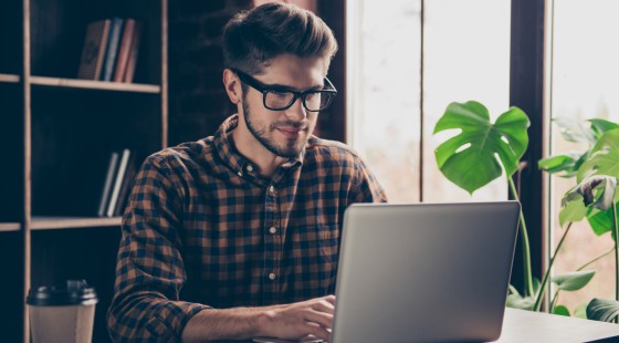 Ein junger Mann sitzt auf einem Schreibtisch und bedient einen Laptop.