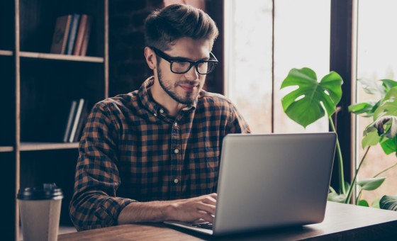 Ein junger Mann sitzt auf einem Schreibtisch und bedient einen Laptop.