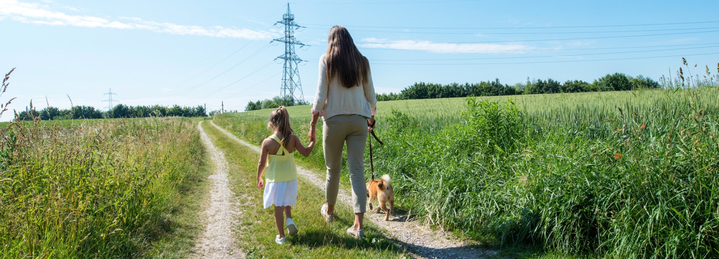 Eine Mutter spaziert mit ihrer kleinen Tochter und einem Hund über einen Feldweg, im Hintergrund eine Stromleitung.